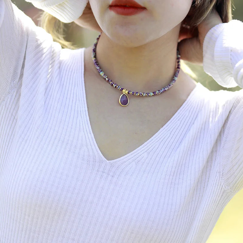 A person wearing a white V-neck top and showcasing a purple Amethyst choker necklace with crystal charms and beads. ALLORA JADE
