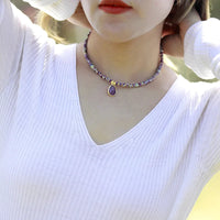 A person wearing a white V-neck top and showcasing a purple Amethyst choker necklace with crystal charms and beads. ALLORA JADE