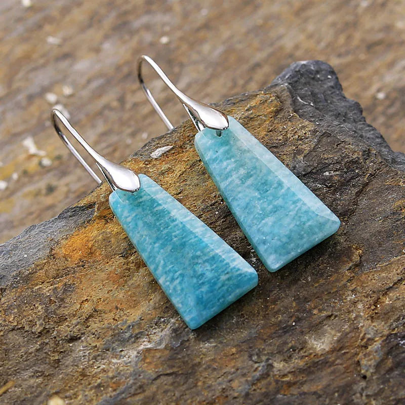 A pair of silver-colored brass earrings with natural Amazonite stones, displayed against a rock background. ALLORA JADE