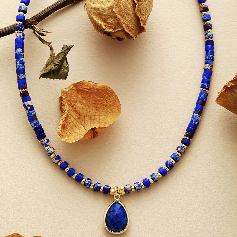 A choker necklace with blue lapis lazuli and jasper beads, featuring a gold-colored crystal charm pendant, displayed against a neutral background with dried leaves. ALLORA JADE
