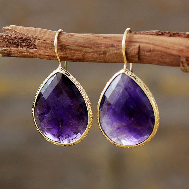 A pair of gold-colored copper earrings with a faceted amethyst centerpiece, displayed on a wooden branch. | ALLORA JADE
