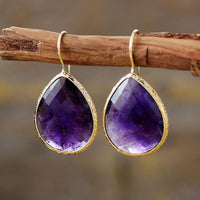 A pair of gold-colored copper earrings with a faceted amethyst centerpiece, displayed on a wooden branch. | ALLORA JADE