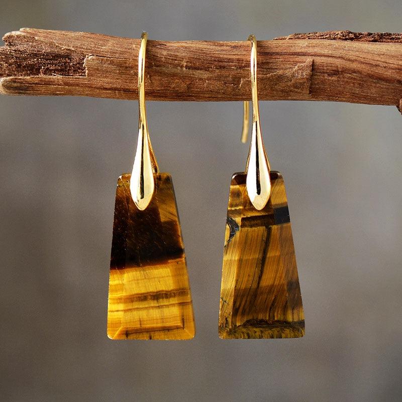 A pair of gold-colored brass earrings with Tiger's Eye stone, displayed hanging on a wooden branch. | ALLORA JADE