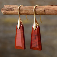 A pair of gold-colored brass drop earrings with red jasper stones, displayed hanging on a wooden branch. ALLORA JADE