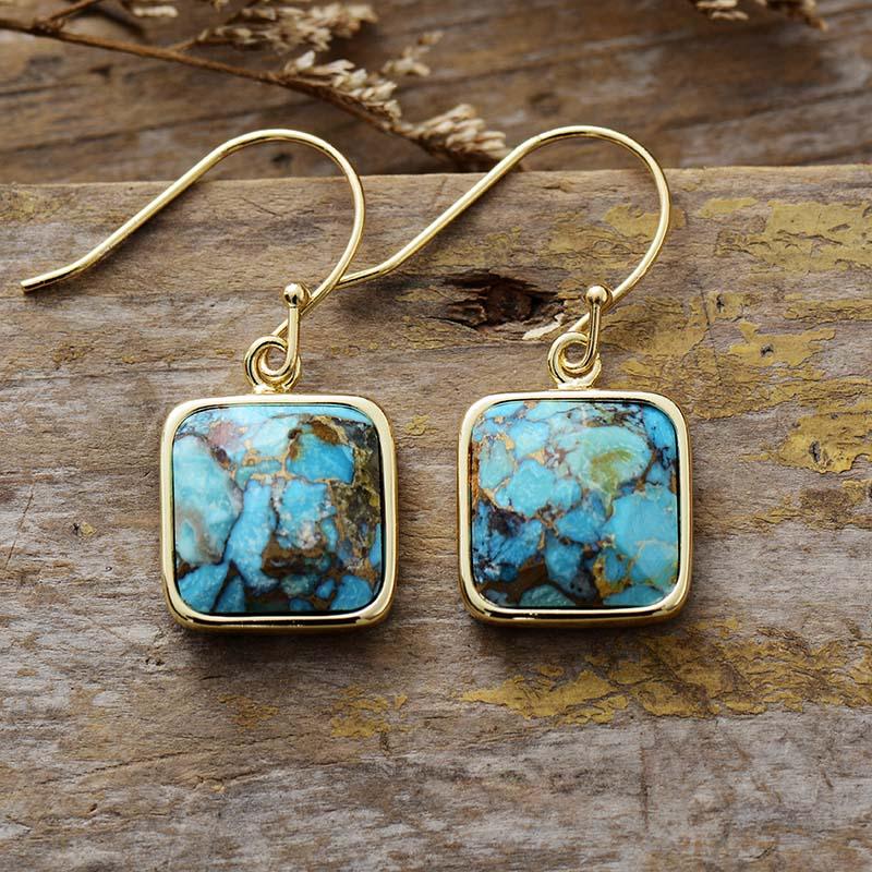 A pair of square turquoise drop earrings with gold-coloured frames, displayed on a wooden surface. ALLORA JADE