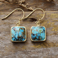 A pair of square turquoise drop earrings with gold-coloured frames, displayed on a wooden surface. ALLORA JADE