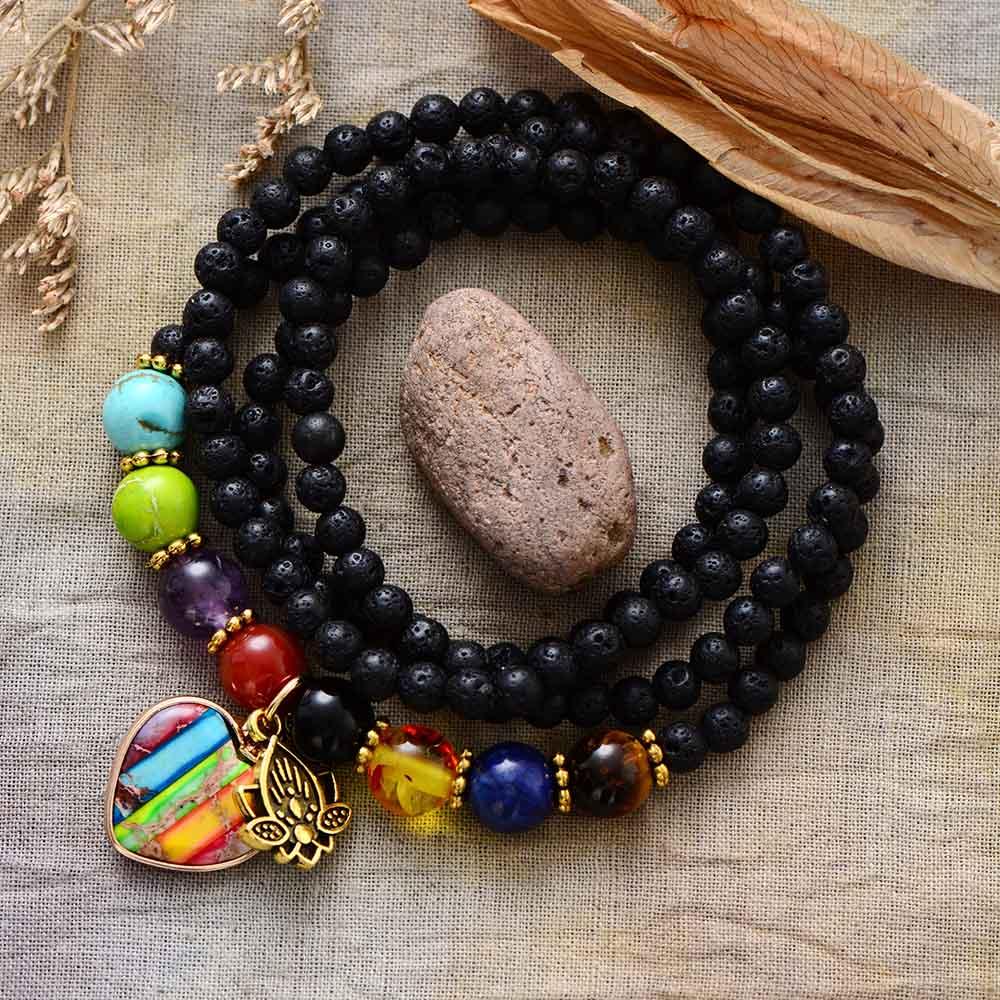 A lava stone bracelet with chakra heart charm beads in various colors including black, along with jasper, amethyst, lapis, turquoise, and agate, displayed on a linen-like surface with a decorative rock in the background. ALLORA JADE