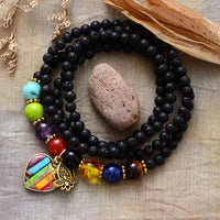 A lava stone bracelet with chakra heart charm beads in various colors including black, along with jasper, amethyst, lapis, turquoise, and agate, displayed on a linen-like surface with a decorative rock in the background. ALLORA JADE