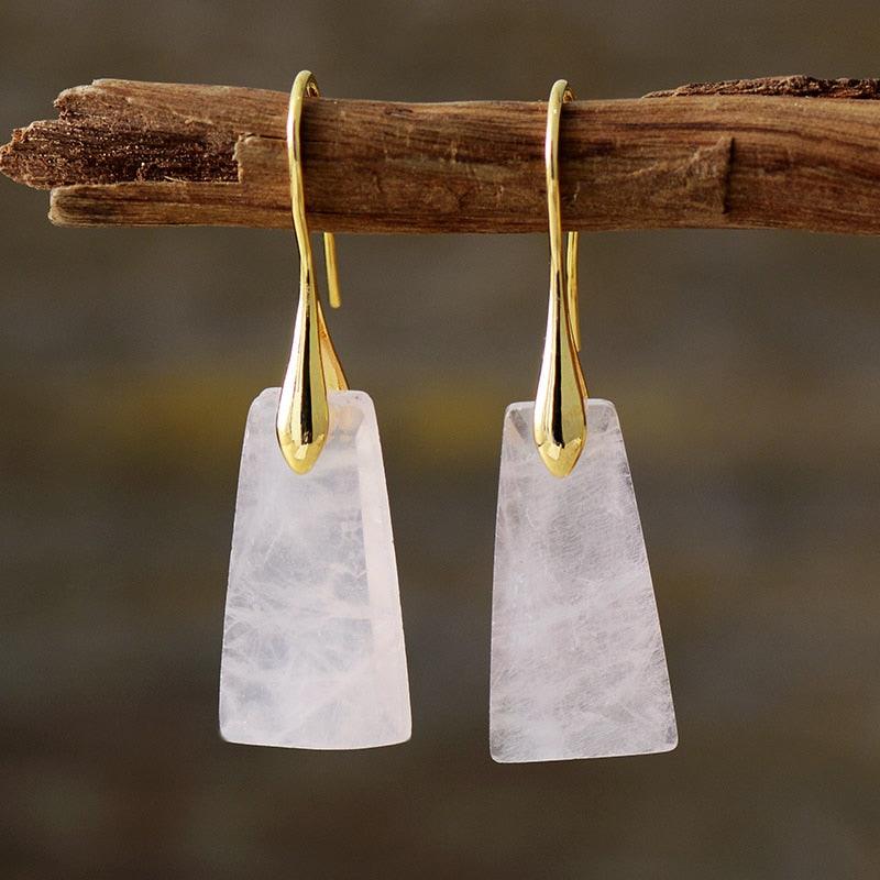 A pair of gold-coloured brass earrings with rose quartz stones, suspended from a branch. ALLORA JADE