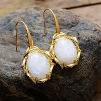 A pair of gold-colored drop earrings featuring round Moonstone stones, displayed on a rock. | ALLORA JADE