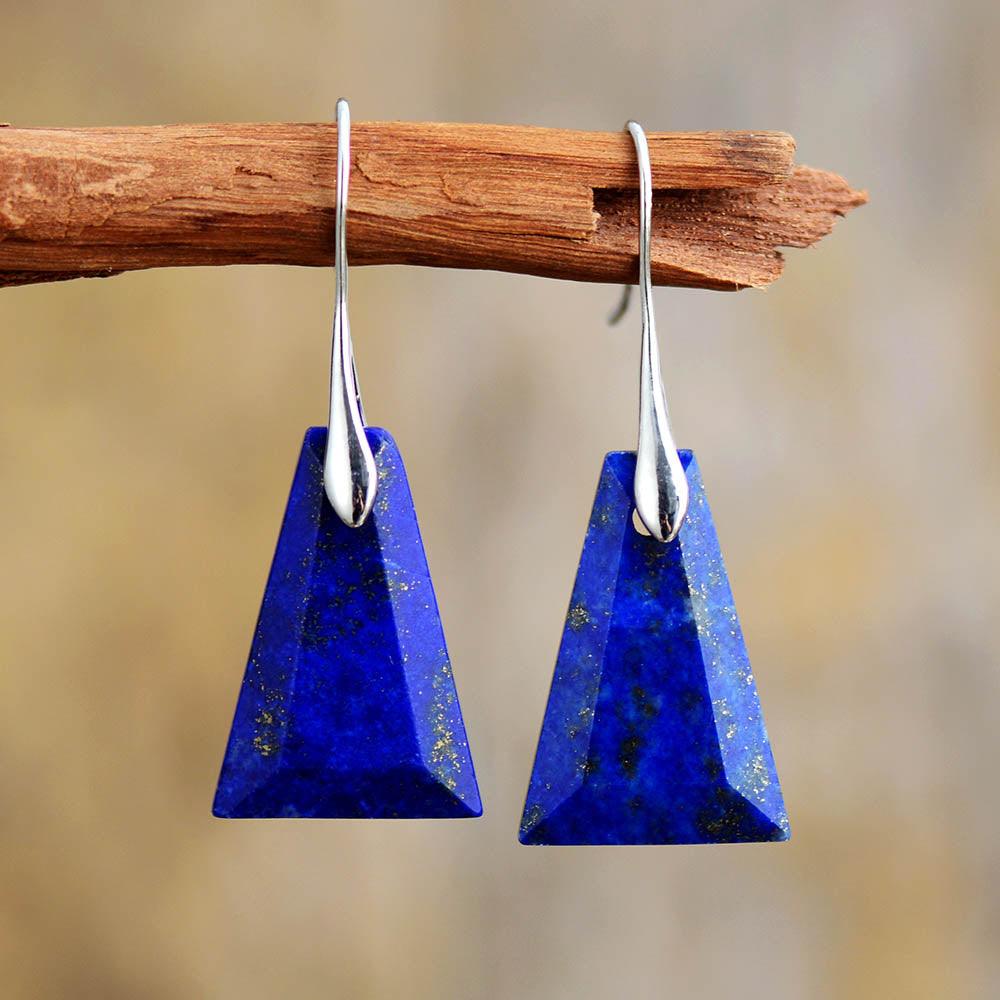 A pair of Lapis Lazuli earrings with trapezoid shape, set in silver-colored brass, displayed hanging on a wooden branch against a blurred background. ALLORA JADE