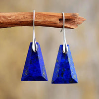 A pair of Lapis Lazuli earrings with trapezoid shape, set in silver-colored brass, displayed hanging on a wooden branch against a blurred background. ALLORA JADE