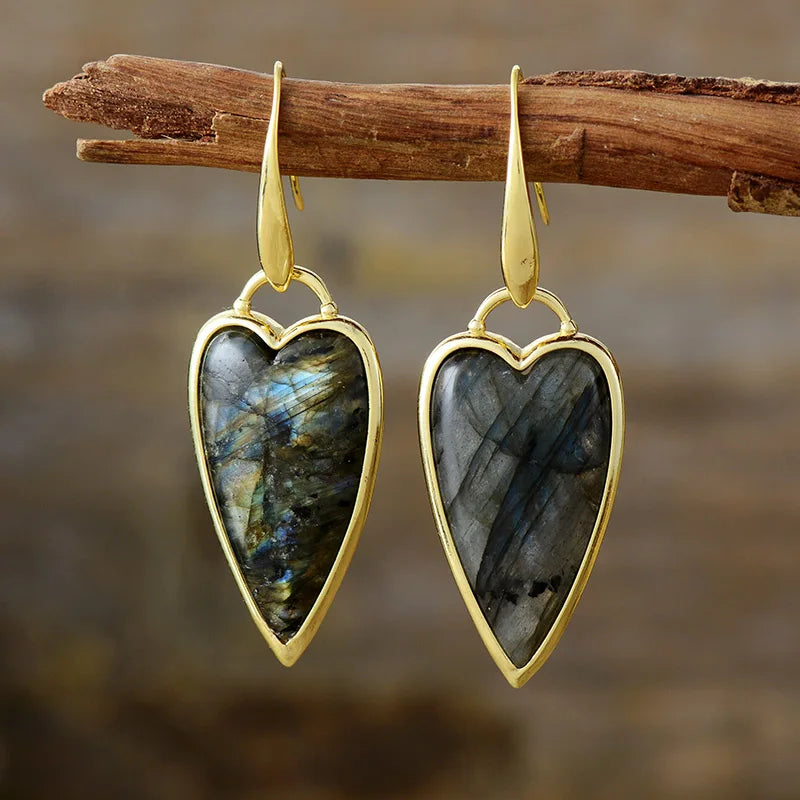 A pair of heart-shaped dangle earrings with polished gold-toned metal borders and hooks, featuring natural labradorite stones with blue and green iridescence, hanging from a rustic wooden branch against a blurred brown background.