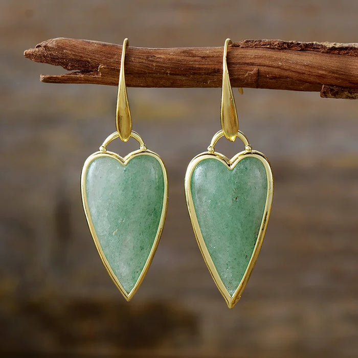 A pair of heart-shaped dangle earrings with polished gold-toned metal borders and hooks, featuring natural green aventurine stones, hanging from a rustic wooden branch against a blurred brown background.