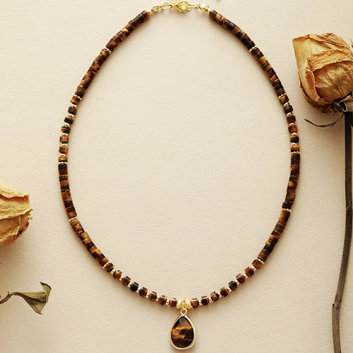 A choker necklace with Tiger's Eye beads and a matching Tiger's Eye charm pendant, displayed on a neutral background with some decorative elements. ALLORA JADE