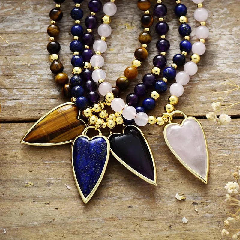 A bohemian-style beaded necklace with four heart-shaped stone pendants in gold settings, displayed on a rustic wooden surface. The necklace features multiple strands of beads including lapis lazuli, rose quartz, tiger's eye, and dark amethyst, accented with small gold beads.