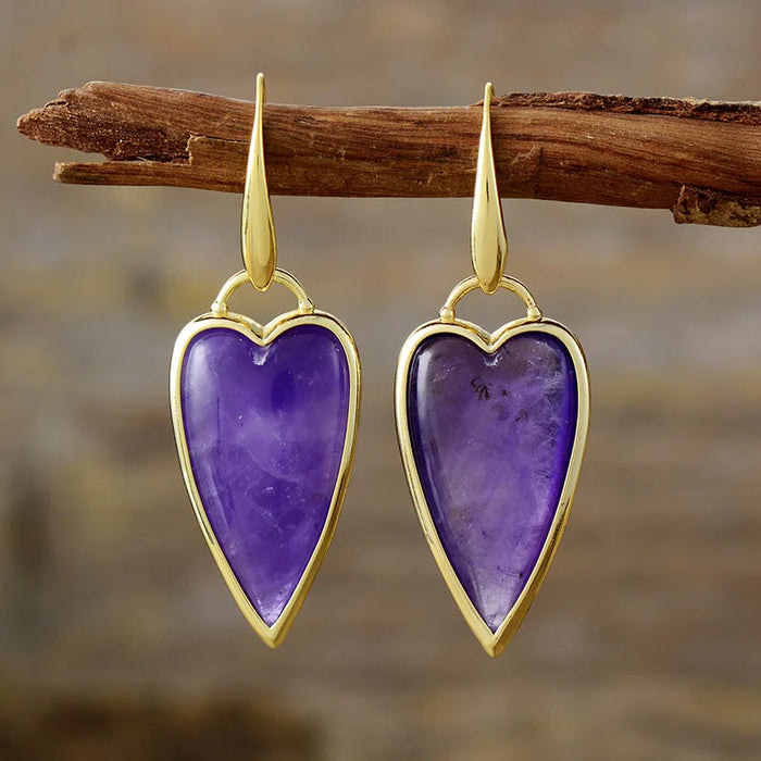 A pair of heart-shaped dangle earrings with polished gold-toned metal borders and hooks, featuring natural amethyst stones, hanging from a rustic wooden branch against a blurred brown background.