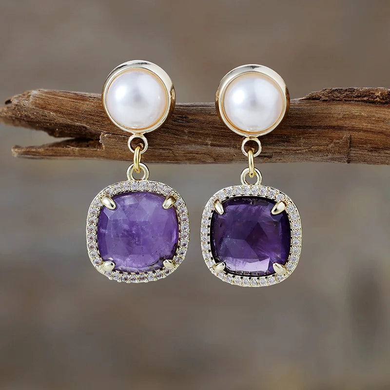 Amethyst Pavé Cushion Dangle Earrings featuring purple gemstone drops framed by sparkling rhinestones, with imitation pearl studs in gold-coloured settings, displayed on rustic wood.