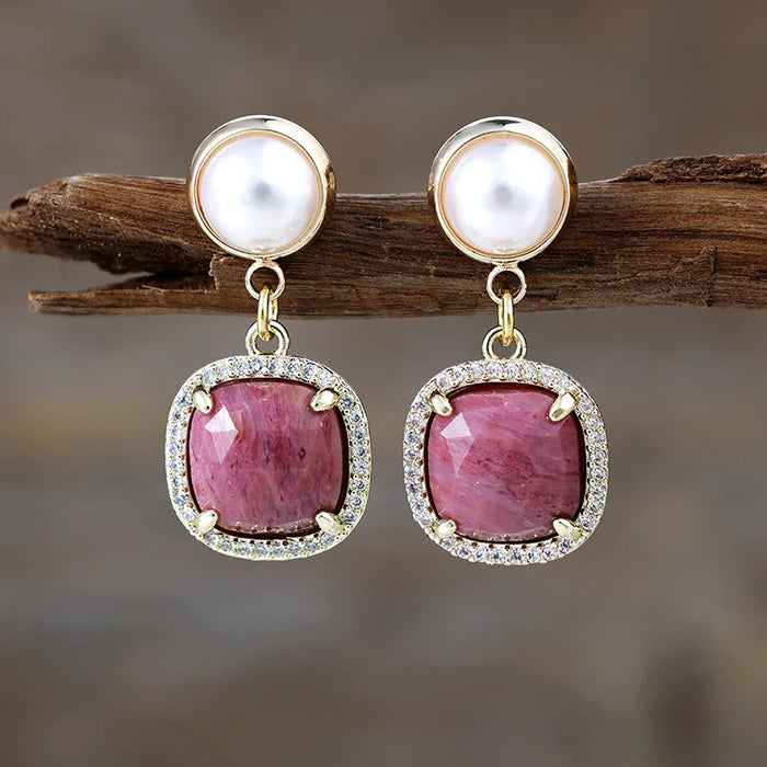 Rhodonite Pavé Cushion Dangle Earrings featuring rosy-pink gemstone drops framed by sparkling rhinestones, with imitation pearl studs in gold-coloured settings, displayed on rustic wood.