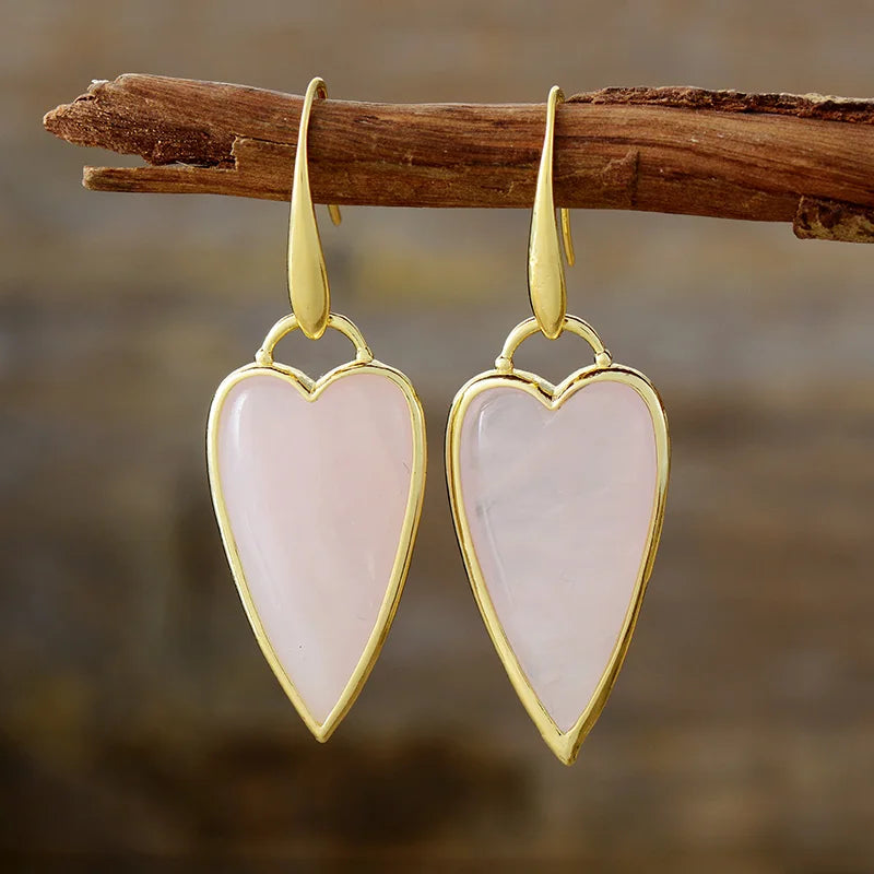 A pair of heart-shaped dangle earrings with polished gold-toned metal borders and hooks, featuring natural rose quartz stones, hanging from a rustic wooden branch against a blurred brown background.