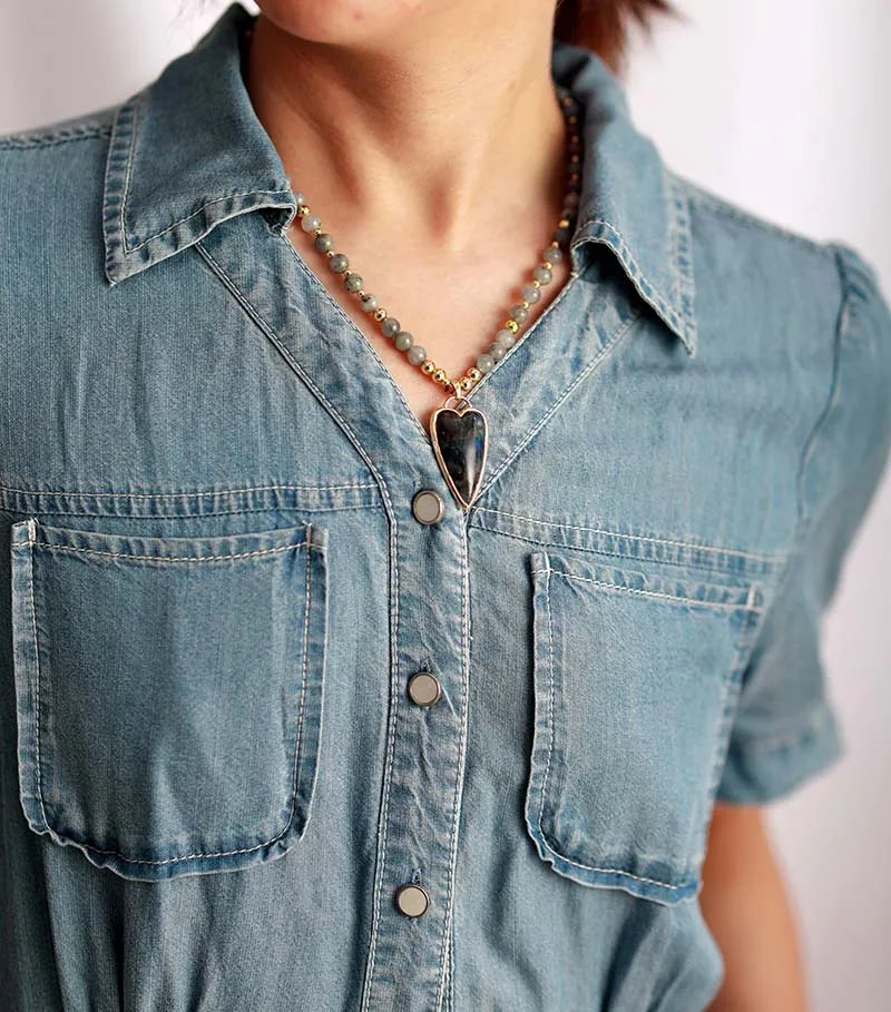 A close-up of a person wearing a light blue denim short-sleeve button-up shirt and a beaded necklace with a dark heart-shaped pendant.