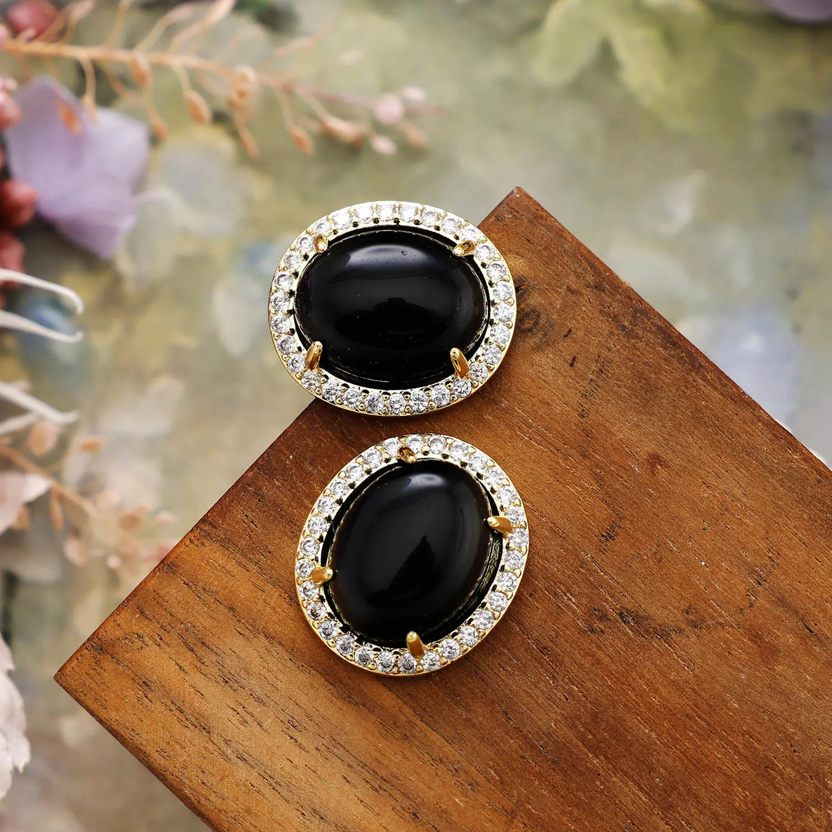 Black Onyx Pavé Oval Stud Earrings featuring polished black gemstones framed by sparkling rhinestones in gold-coloured settings, displayed on rustic wood.