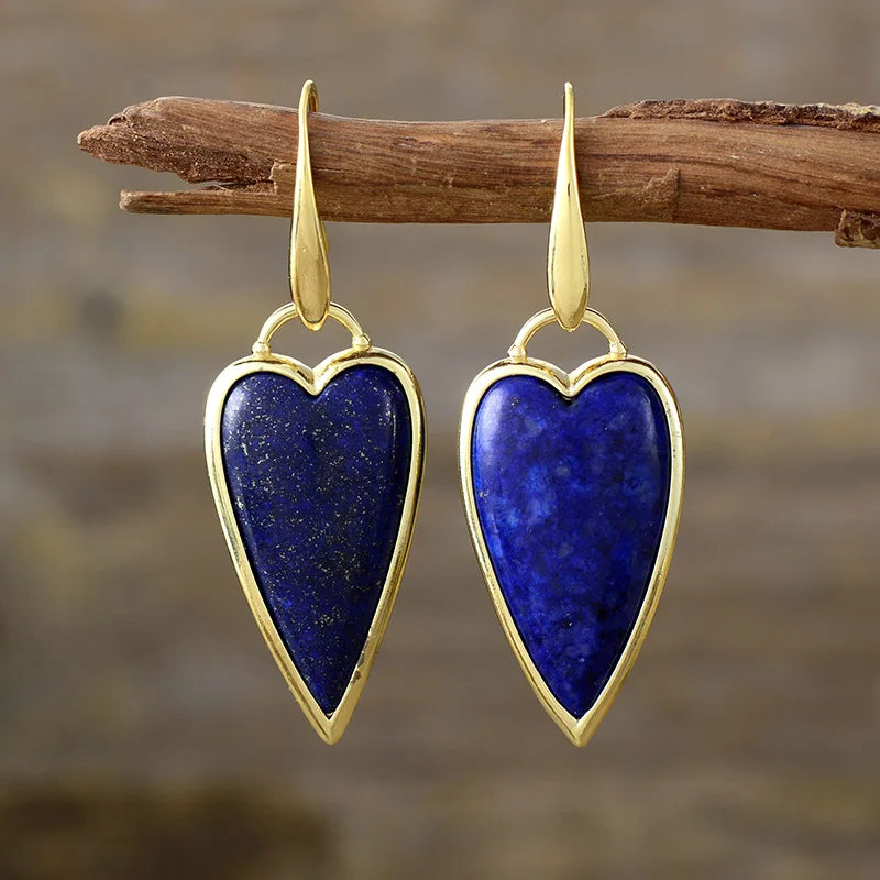 A pair of heart-shaped dangle earrings with polished gold-toned metal borders and hooks, featuring natural lapis lazuli stones, hanging from a rustic wooden branch against a blurred brown background.