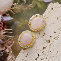 Rose Quartz Pavé Oval Stud Earrings featuring polished pink gemstones framed by sparkling rhinestones in gold-coloured settings, displayed on stone surface with floral background.