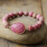 A stretchy bracelet with 8mm round Rhodonite beads and a faceted Rhodonite charm, displayed on a stone surface.