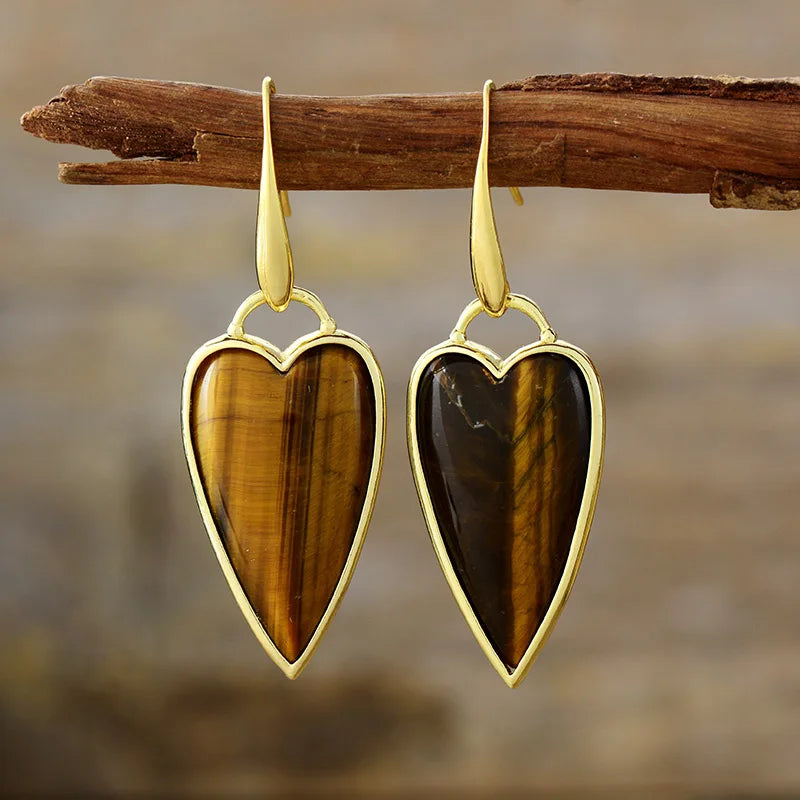 A pair of heart-shaped dangle earrings with polished gold-toned metal borders and hooks, featuring natural Tiger's Eye stones, hanging from a rustic wooden branch against a blurred brown background.